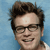 Pixelated close-up of a person with dark brown hair, wearing black-framed glasses, against a light blue background.