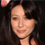 A pixelated close-up of a person with dark hair and light skin, wearing a dark top, against a dark background with a faint pinkish glow in the upper left corner.