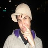 A person wearing a light-colored beanie and a gray jacket with purple collar accents holds a finger to their lips against a dark background with faint lights.