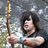 A person with dark hair holds a wooden staff, standing before a blurred background of foliage and stone.