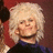 Pixelated portrait of a person with voluminous light-colored hair, wearing dark clothing and a red shoulder accent against a brown background.