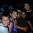 Group of people in dark clothing with one person in blue top, smiling faces, dark background, and a light gray area on the right.