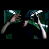 A person in a dark hoodie and green shirt holds a camera, transitioning from a raised position to a lowered position