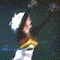 Animated sequence of a person with curly blonde hair performing on stage, wearing a black top with red and yellow stripes across the chest, against dark and blue backgrounds.