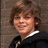 A pixelated image of a person with light brown hair wearing a dark jacket over a white-collared shirt, next to blue pixelated text on a black background.