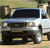 Static image of a white van with dark windows and black front bumper, positioned on a light brown surface with green and brown background elements.