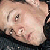 Pixelated close-up of a person's face with light skin tone, dark hair, and visible eyes and nose against a dark background.
