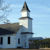 Pixelated static image of a light blue building with a tall steeple under a clear sky, featuring a brown roof and dark windows.