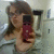 A person with brown hair holds a red smartphone in a bathroom mirror selfie, wearing a light gray top.