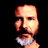 Pixelated static image of a bald man's face and upper neck with light skin tone, visible facial features including eyes, nose, mouth, and beard stubble, wearing a white collared shirt against black background.