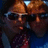 Two figures in dark clothing with brown hair under a warm light source, one figure's face partially obscured by a dark shape.