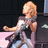 Pixelated image of a person with long blonde hair wearing dark clothing and light-colored pants, seated against a dark background.