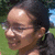 A person with dark hair tied back, wearing glasses, smiling outdoors with green grass and blue sky in the background.