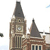 A pixelated static image of a tall clock tower with a dark spire, brown stone facade, and visible clock face, set against a light sky with faint clouds.