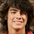 A person with voluminous dark curly hair and a red shirt smiles in three showing slight head movement