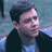 Pixelated static image of a person with dark hair wearing a dark jacket over a light blue shirt against a blurred background.