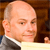 A pixelated close-up of a person with a balding head wearing a dark suit and white shirt, set against a brown wooden background.
