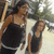 Two figures in dark sleeveless tops stand side-by-side against a blurred background with light and dark tones.