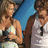 A woman in a white halter top and light blue skirt holds a black camera, standing beside a man in a white tank top with dark hair against a bright blue background.