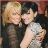Two women embrace in a close-up pose, one with blonde hair in a black mesh top, the other with dark hair in a silver patterned sleeveless top, both smiling against a blurred background.