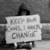 Black-and-white image of a person holding a sign reading "FOOTBALL CHAMPIONS I WANT CHANGE" with a blurred background.