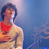 A person with curly dark hair wears a red undershirt under a light gray long-sleeve shirt, standing against a blue-lit background with a microphone and stage equipment visible.