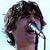 Pixelated close-up of a person with dark curly hair holding a microphone, eyes closed against bright light reflection.