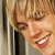 Pixelated close-up of a smiling person with light brown hair and visible teeth, wearing dark clothing against a neutral background.