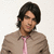 Pixelated static image of a person with dark hair wearing a light brown jacket over a pink shirt against a plain background.