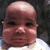 Pixelated close-up of a person's face with dark eyes, pinkish skin tone, and light-colored clothing against a dark background.
