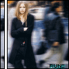 A woman with long blonde hair in a black coat stands against a blurred urban background, arms crossed.