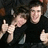 Two people in dark clothing pose closely together on the left side of a purple background with yellow pixelated text.
