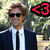 Pixelated image of a person in a dark suit and white shirt with a black tie, wearing dark sunglasses, standing against a blurred green and beige background with a red and black symbol on the right.