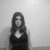 A grayscale image of a person with long hair wearing a dark sleeveless top, standing against a plain wall background.