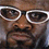 Pixelated close-up of a person's face wearing large white-framed glasses with dark lenses, brown skin tone, and visible facial features including nose and mouth area.