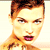 Pixelated close-up of a person with short brown hair, light skin, and red lips, wearing a light-colored top against a pale background.