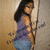 A woman with long dark hair in a purple sleeveless top and blue jeans stands against a light brown wall, holding a thin vertical object.