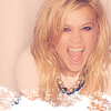 Pixelated close-up of a person with long wavy blonde hair wearing a white top and dark beaded necklace against a light background.