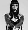 Black-and-white image of a person with long dark hair and bangs wearing a black lace top, crossed arms, and visible chest area.