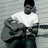 Pixelated image of a person in white shirt and dark pants holding an acoustic guitar, with overlaid pixelated text blocks on light background.