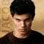Pixelated portrait of a person with short dark hair wearing a dark shirt against a light brown textured background.