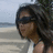 Pixelated static image of a person with long dark hair wearing sunglasses and a white sleeveless top, standing against a blurred beach background.