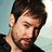A man with dark hair and a beard wears a black shirt against a light blue background, his head slightly tilted downward.