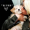 A person in a dark green jacket holds a small light brown dog with closed eyes, while a blonde-haired person kisses the dog's head.