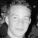 Black-and-white pixelated image of a person with short curly hair, neutral expression, and dark clothing against a blurred background.