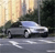 Pixelated static image of a silver sedan on a road with blurred background trees and buildings.
