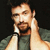 Pixelated image of a person with short brown hair and a beard, wearing a dark green shirt, holding a white object near their face against a black background.