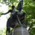 Pixelated image of a dark statue on a pedestal with green foliage background, showing a figure holding a long object and another figure standing beside it.