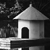 Black-and-white pixelated image of a small house with a triangular roof, single dark door, and light walls, set against a dark background with faint tree-like shapes.