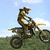 A rider on a dirt bike in mid-air against a blue sky with clouds and green grass background.